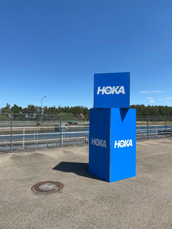 Totem rigide HOKA en zone bord de canal — signalétique premium pour les espaces à forte exposition photographique. Totem rigide HOKA — zone canal, fond de parcours triathlon